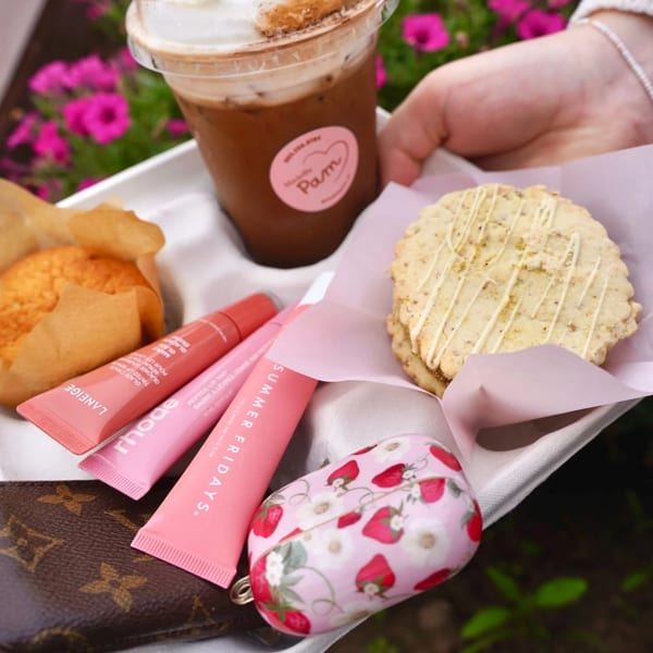 Hand holding a tray with iced coffee, muffin, cookie, lip balms, and a strawberry-patterned case.