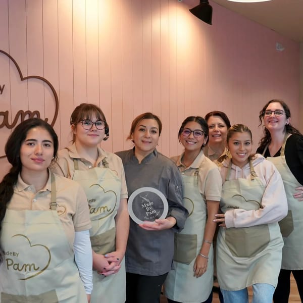 Seven smiling women in aprons stand in front of a pink wall with the "MadeBy Pam" logo.