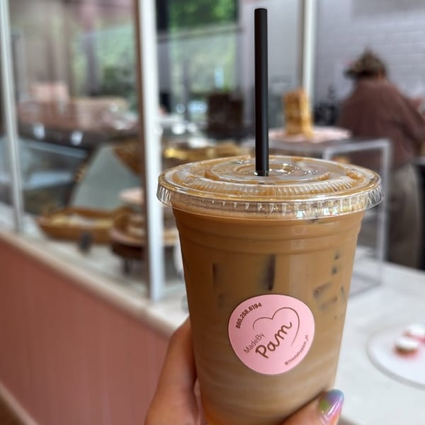 Hand holding an iced coffee with a pink "MadeByPam" sticker, inside a bakery.
