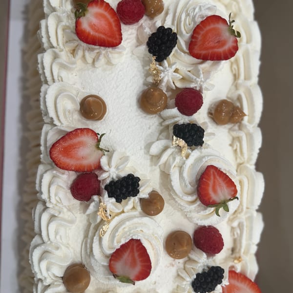 A rectangular cake topped with white frosting, strawberries, raspberries, blackberries, and dulce de leche swirls.