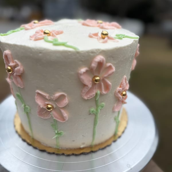 White frosted cake decorated with pink buttercream flowers, green stems, and gold pearl centers.