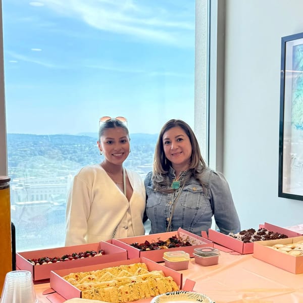 Two women smiling behind a table laden with desserts in pink boxes, with a city view outside.