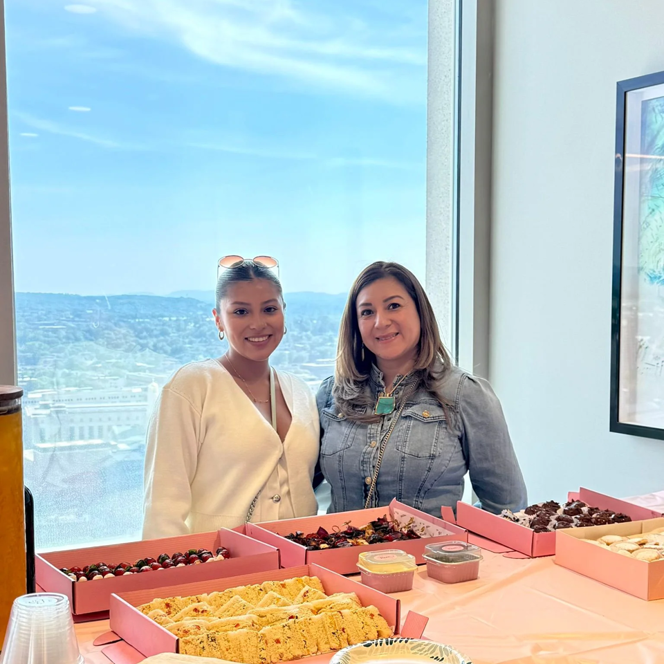 Two women smiling behind a table laden with desserts in pink boxes, with a city view outside.