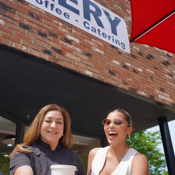 Two smiling women, one holding a coffee cup, in front of a bakery sign and brick wall.