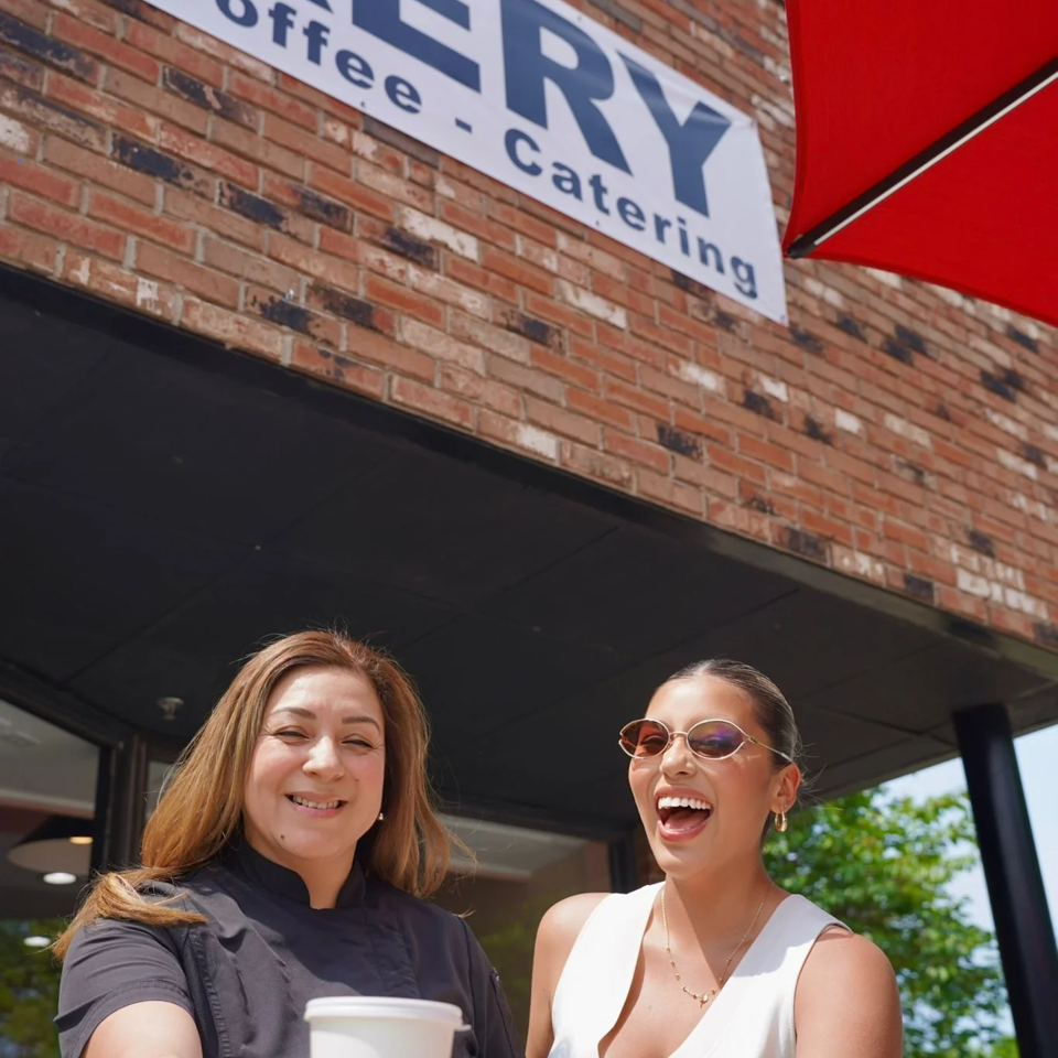 Two smiling women, one holding a coffee cup, in front of a bakery sign and brick wall.