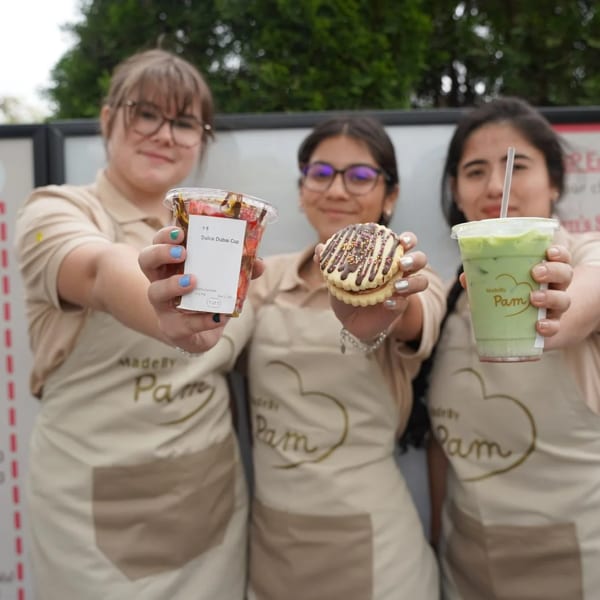 Three MadeBy Pam employees in aprons hold up a Dulce Dubai Cup, a cookie, and a green drink.
