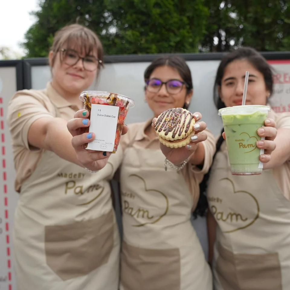 Three MadeBy Pam employees in aprons hold up a Dulce Dubai Cup, a cookie, and a green drink.