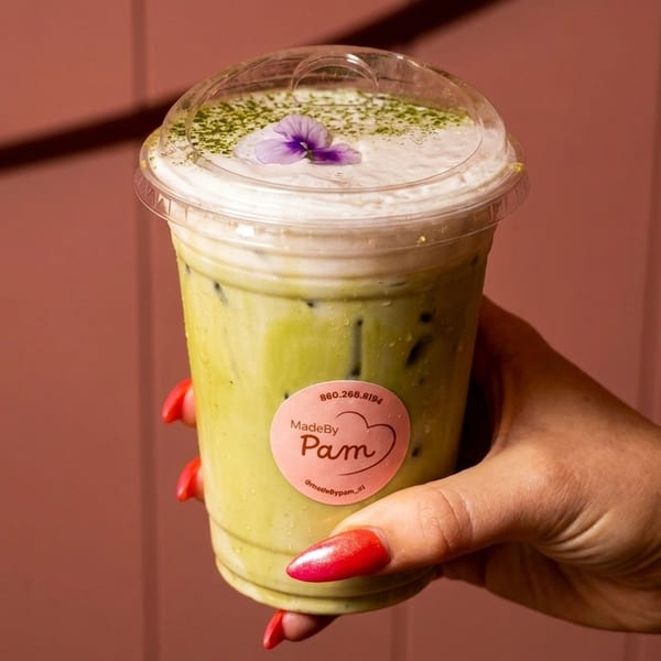 A hand with red nails holds a MadeByPam iced matcha latte with foam and a purple flower.