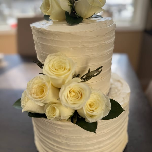 Two-tiered white wedding cake with textured frosting, adorned with white roses and green leaves.