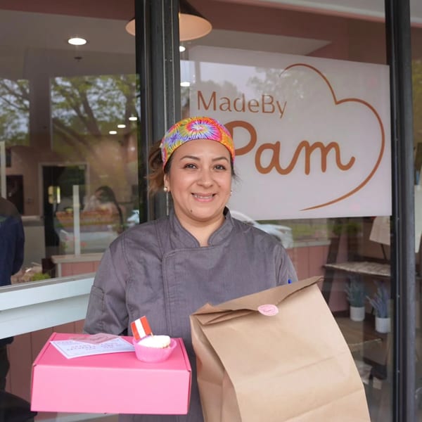 A smiling woman in a chef's coat holds a pink box and a brown bag outside "MadeBy Pam".