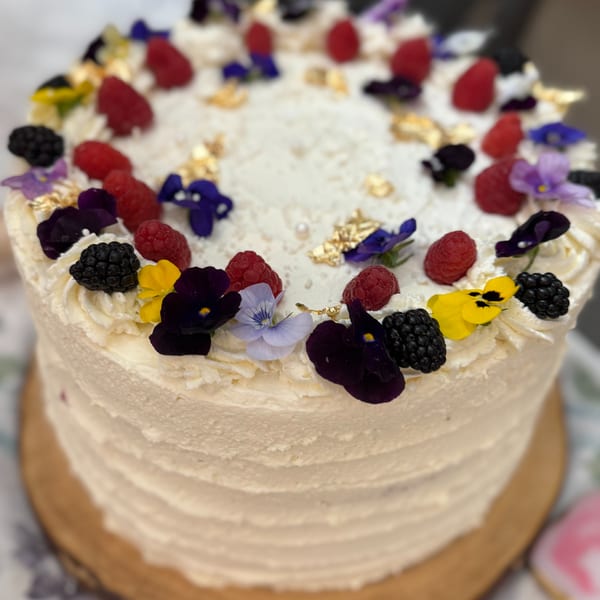 White frosted cake adorned with raspberries, blackberries, edible flowers, and gold flakes on a wooden stand.